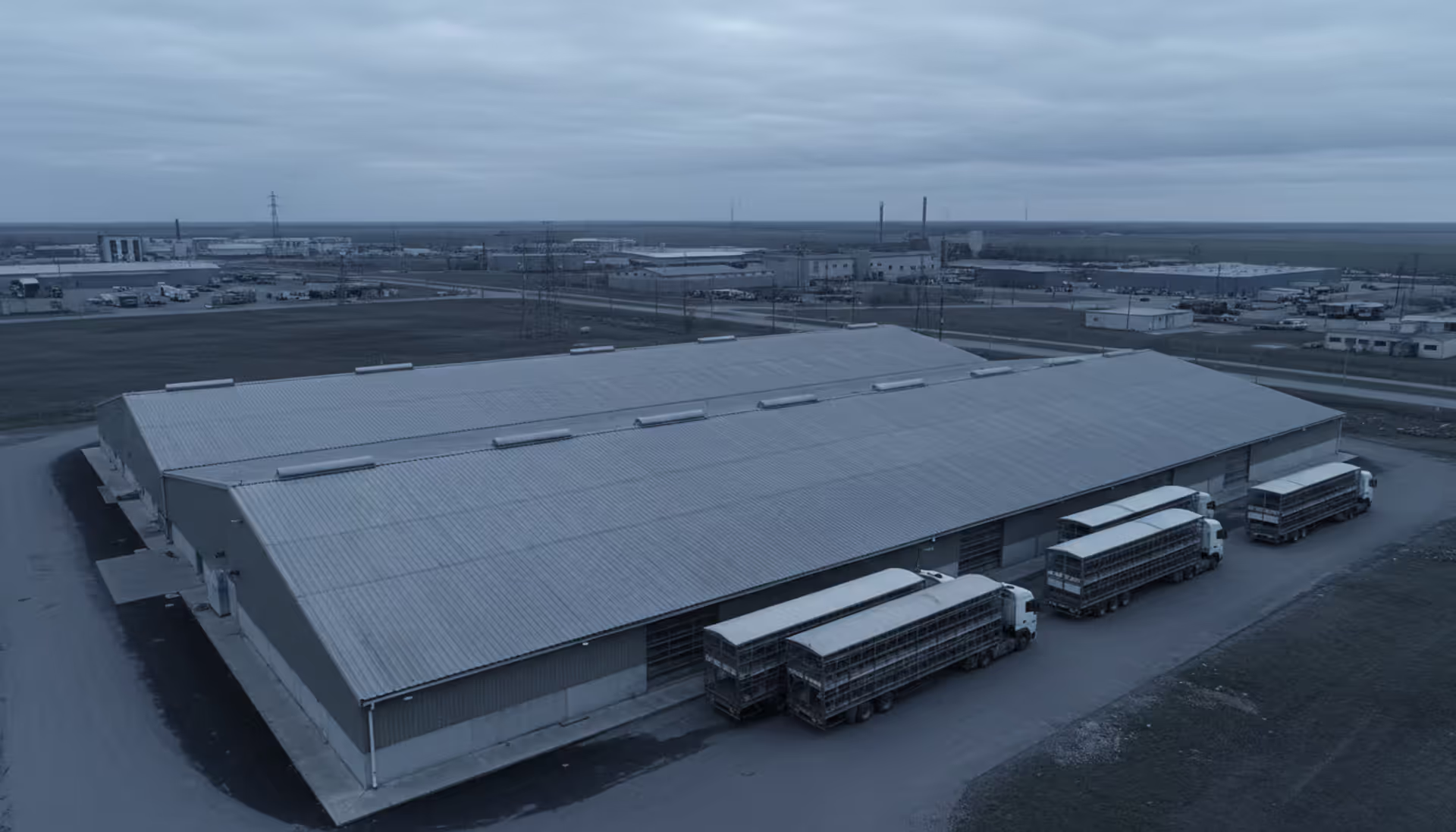 Aerial view of a large industrial poultry barn with metal roof and transport trucks parked outside under an overcast sky